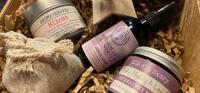 w pudełku z karbowanego kartonu wyłożonego kartonowymi ścinkami leżą 3 kosmetyki w fioletowych opakowaniach: sos dla skóry słodkie migdały i lawenda, hydrolat lawendowy, aromaterapia miłość drzewo różane.